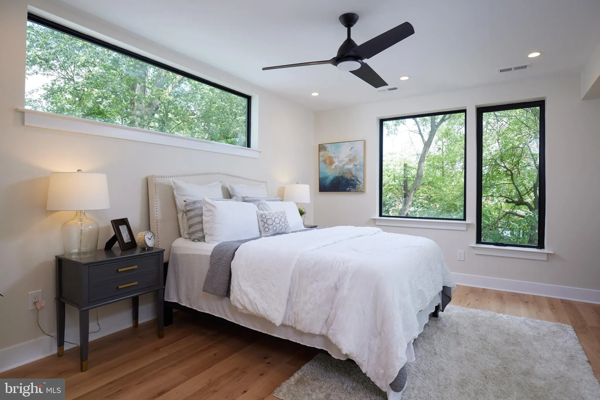Primary bedroom with white bedding, black nightstand, large windows with tree views